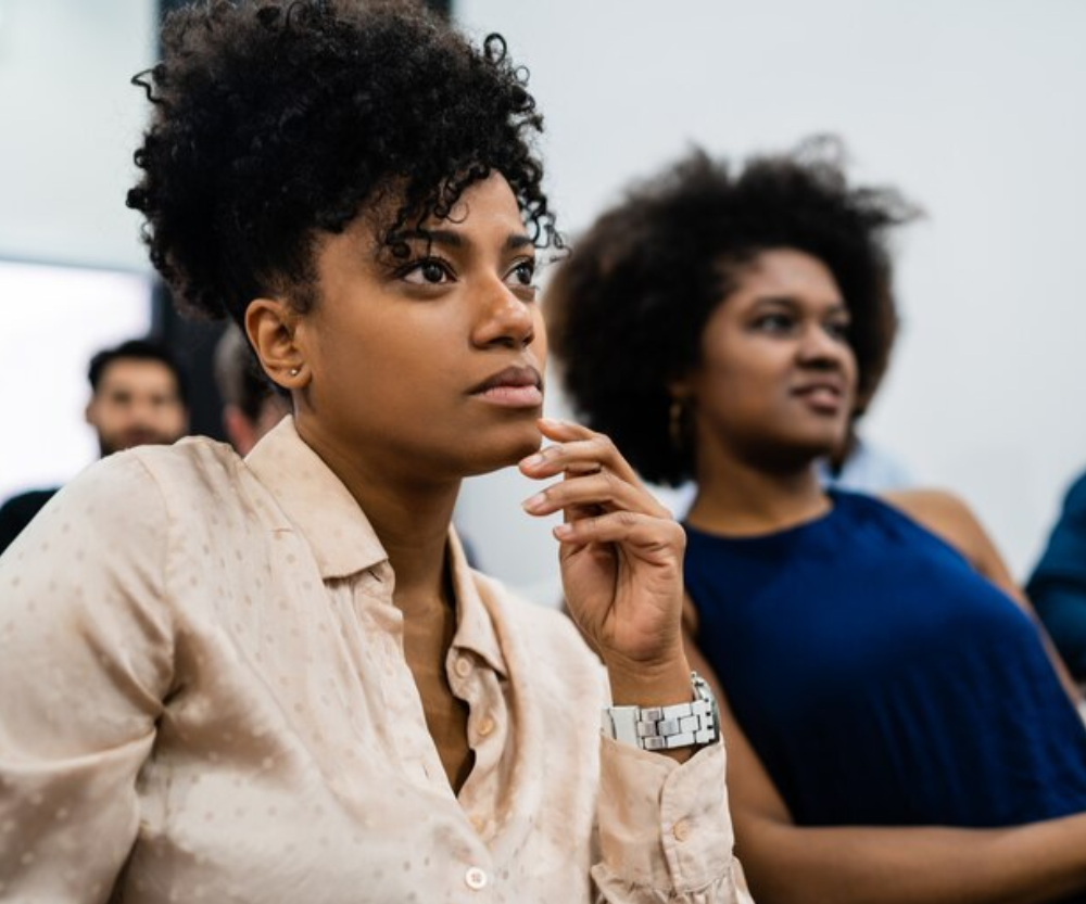 Foto de duas mulheres negras em um ambiente de sala de aula ou conferência, com expressões de concentração e interesse. A mulher em primeiro plano veste uma camisa bege e tem cabelo cacheado preso em um coque. A imagem representa diversidade, inclusão e está associada à heteroidentificação.